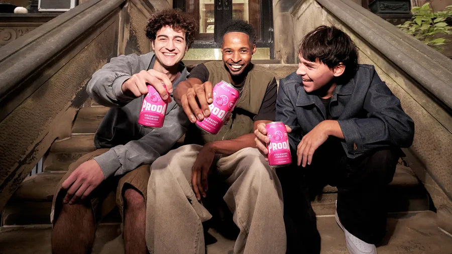 Three friends on a brownstone stoop raise cans of PRODA Strawberry Lemonade.