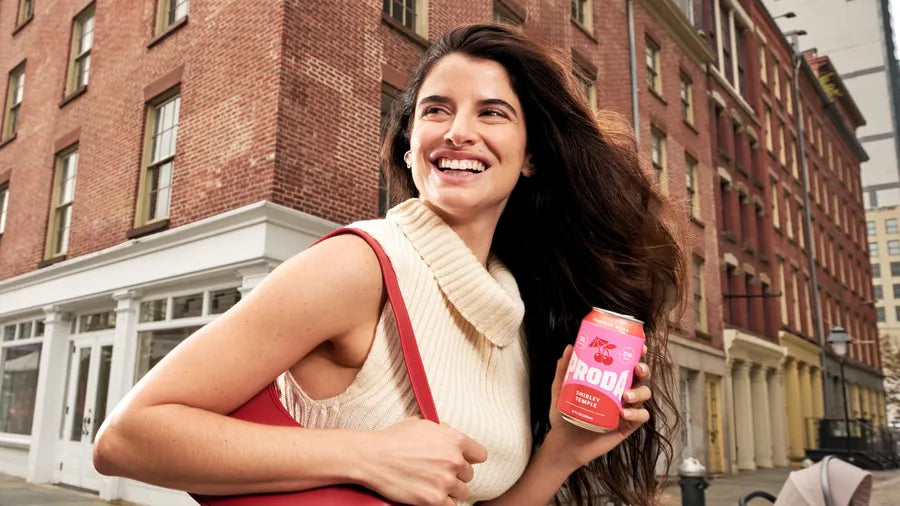 A woman smiles on a city street holding a can of PRODA Shirley Temple.