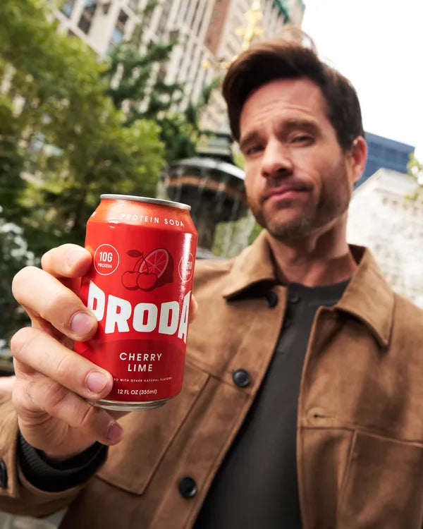 A man in a city park holds a can of PRODA Cherry Lime out toward the camera.