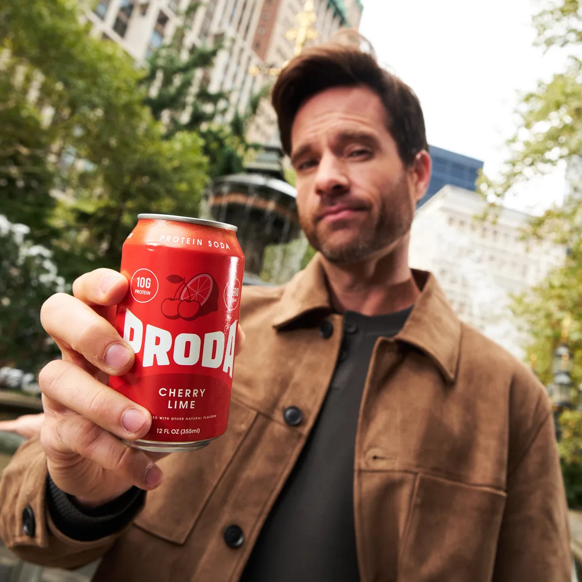 A man in a suede jacket holding a can of PRODA Cherry Lime toward the camera in a New York park