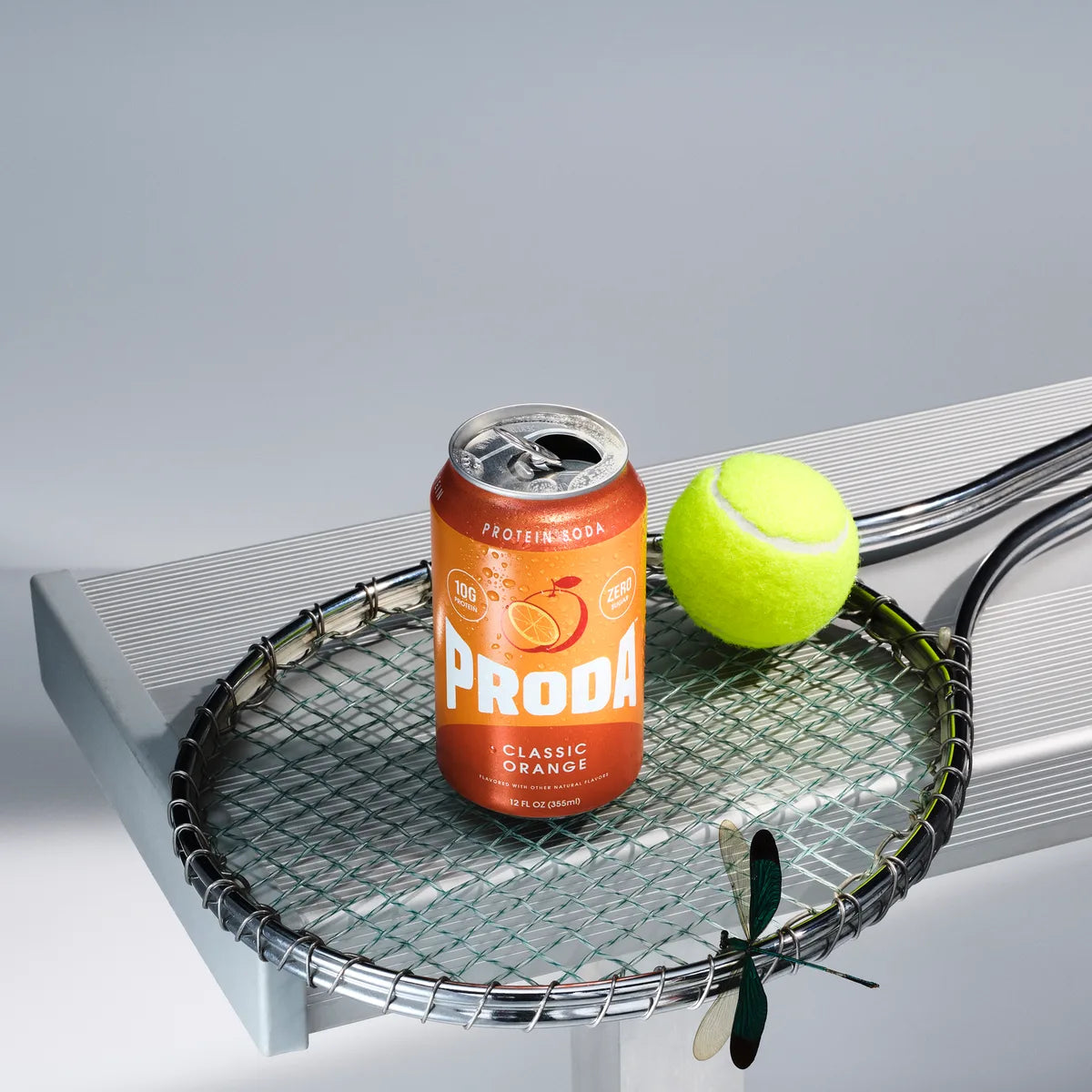 A can of PRODA Classic Orange resting on a tennis racket beside a tennis ball