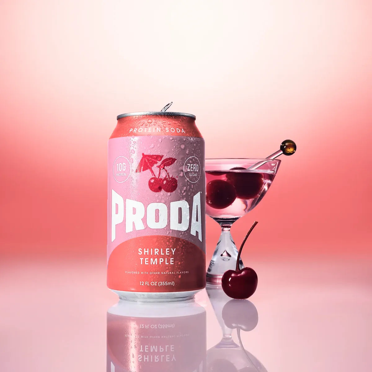 A can of PRODA Shirley Temple beside a cocktail glass with a cherry on a pink backdrop
